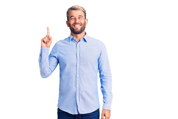 Mladý pohledný blonďatý muž v elegantní košili ukazuje a ukazuje prstem číslo jedna a zároveň se usmívá sebevědomě a šťastně.  - Fotografie, Obrázek