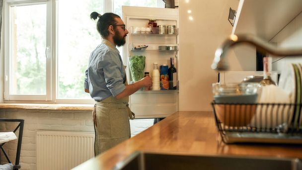 Young man, professional cook in apron taking ingredients out of the fridge while getting ready to prepare a meal, standing in the kitchen. Cooking at home concept - Foto, Bild