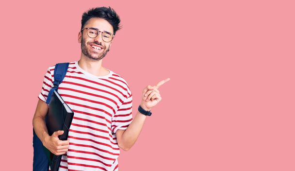 Joven hombre hispano con mochila de estudiante y gafas sosteniendo aglutinante sonriendo feliz señalando con la mano y el dedo hacia un lado  - Foto, imagen