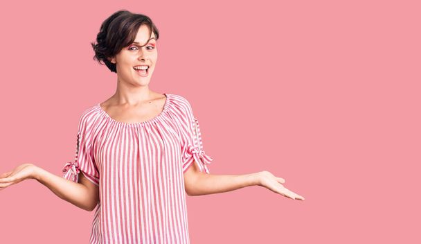 Mooie jonge vrouw met kort haar dragen casual zomerkleding glimlachen tonen beide handen open palmen, presenteren en reclame vergelijking en evenwicht  - Foto, afbeelding
