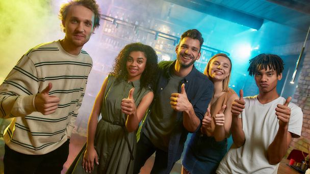 Young men and women smiling, showing thumbs up while posing together for camera. Multiracial group of friends hanging out at party in the bar - Photo, Image