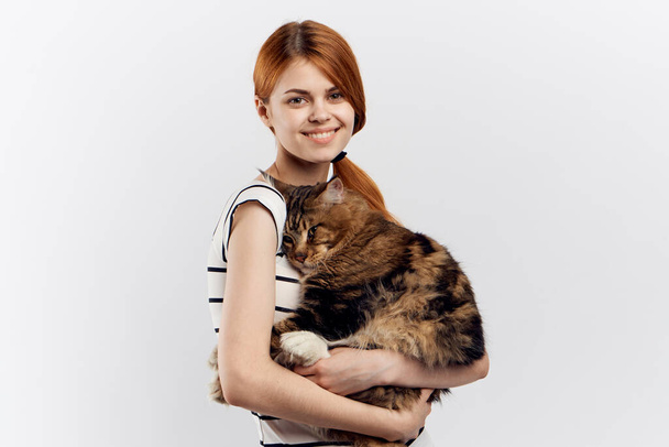 Red-haired woman holds in her hand a cat striped T-shirt light background home pet - Photo, Image