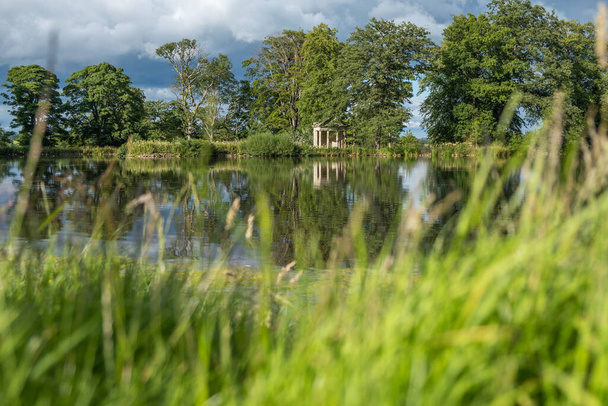 Вид з розмитою травою на передньому плані через красиве озеро з відображенням дерев у бік старої покинутої споруди посеред сільської сільської місцевості
 - Фото, зображення