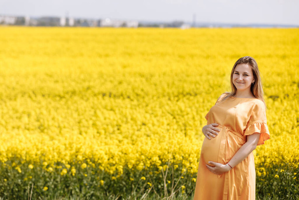 Mladá těhotná žena ve žluté řepce pole drží její břicho a na sobě žluté šaty na slunné letní den. - Fotografie, Obrázek