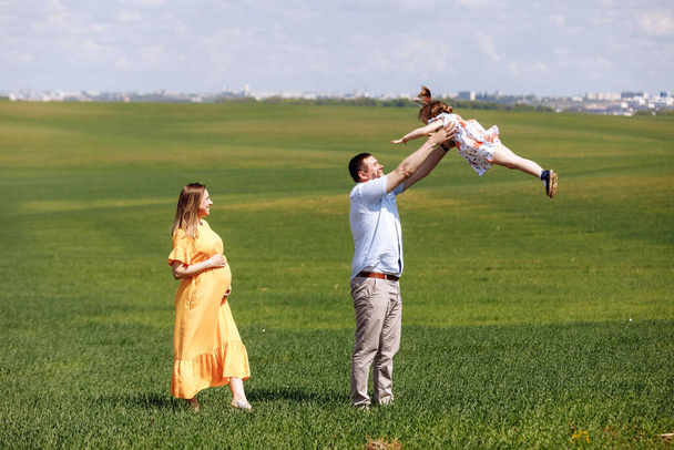 Happy pregnant family with little daughter spending time together in sunny green field on summer day. - Photo, image