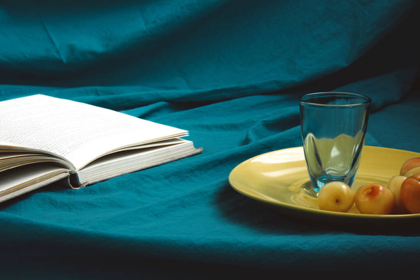 Cereza amarilla dulce y un vaso de precipitados se encuentran en un plato amarillo, junto a un libro abierto. Fondo de tela turquesa. Fotografía de comida oscura. Enfoque selectivo - Foto, imagen