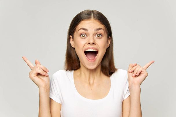 Fröhliche Frau im weißen T-Shirt zeigt Finger zur Seite und weit geöffneten Mund - Foto, Bild
