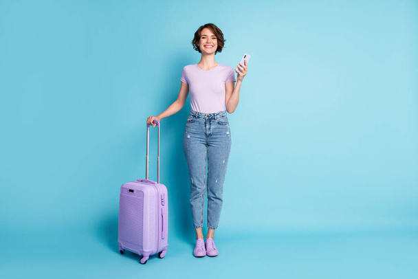 Full size photo of positive cheerful girl tourist hold baggage use smartphone booking taxi hotel wear lilac violet t-shirt denim jeans isolated blue color background - Foto, Bild