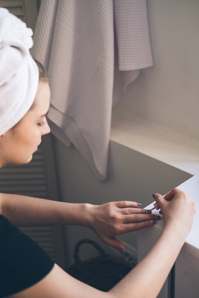 Young beautiful woman with towel on her head after shower in bathroom applies nail polish. The concept of female morning makeup procedures. - Photo, Image