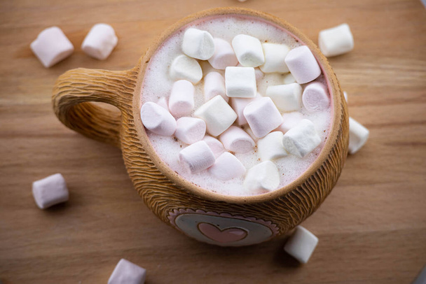 Brown ceramic mug with pink heart. Hot cappuccino coffee and airy pink marshmallows are poured in a cup. On a wooden background. View from above. - Фото, зображення