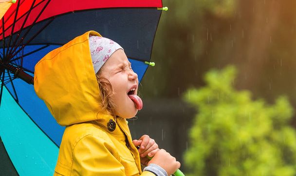 Lustiges kleines Mädchen im gelben wasserdichten Mantel. Kindergesicht mit offenem Mund, der Tropfen auffängt. Kind mit buntem Regenschirm spielt im Garten im Herbstregen - Foto, Bild