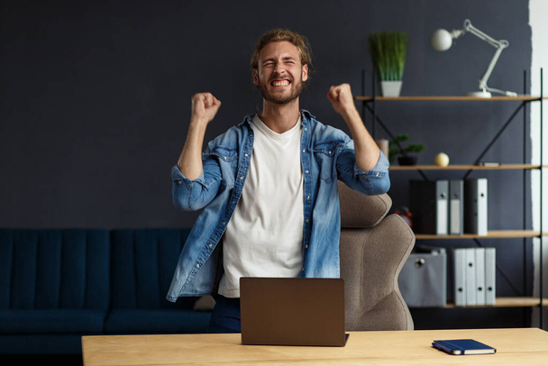 Young handsome long haired curly man working in office with laptop and expressing positivity. Happy businessman got positive news.Rewarded,new contract,good deal, successful negotiation,best day ever - Fotoğraf, Görsel