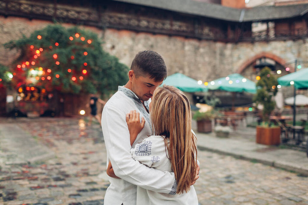 Verliebtes Paar, das in traditionellen ukrainischen Hemden durch die Altstadt von Lwiw spaziert. Menschen lächeln und umarmen sich entspannt vor Arsenal-Café im Regen - Foto, Bild