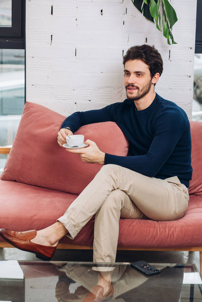positive young man in stylish clothes having breakfast while sitting on the sofa - Photo, image