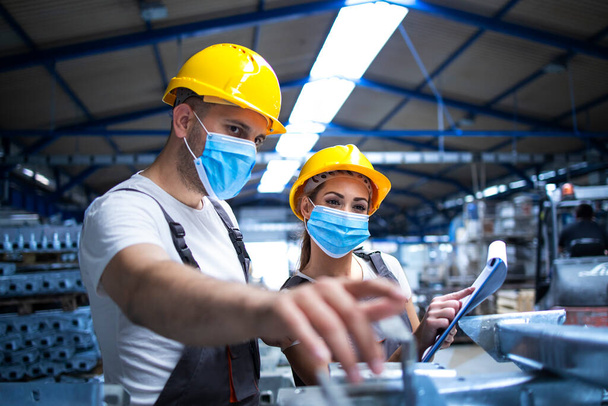 Pracownicy przemysłowi z maskami chronionymi przed wirusem korony rozmawiający o metalowych częściach w fabryce. Osoby pracujące podczas pandemii COVID-19. - Zdjęcie, obraz