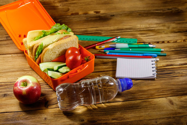 Back to school concept. School supplies, bottle of water, apple and lunch box with sandwiches and fresh vegetables on a wooden desk - 写真・画像