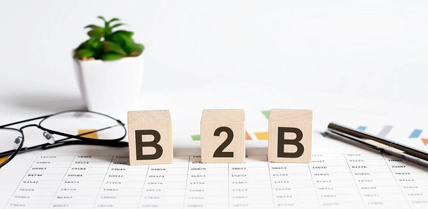 B2B szó koncepció írva fa blokkok, kockák egy könnyű asztal virággal, toll és szemüveg chart háttér - Fotó, kép