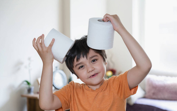 Szczęśliwy młody chłopiec z uśmiechniętą twarzą bawiący się papierem toaletowym, dziecko wkładające oilet roll na głowę, dziecko trzymające dwie białe tkanki, koncepcja opieki zdrowotnej dla dzieci - Zdjęcie, obraz