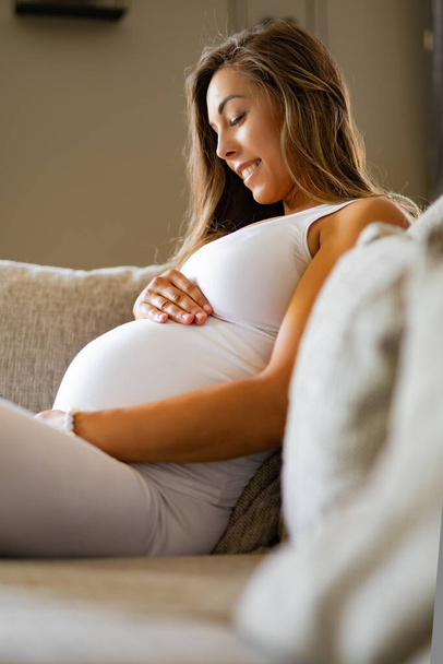 Χαμογελώντας έγκυος γυναίκα κάθεται στον καναπέ κοιτάζοντας το στομάχι της - Φωτογραφία, εικόνα