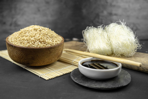bruine rijst in een houten kom, Aziatische keuken achtergrond met bamboe mat, eetstokjes, sojasaus, gember en glas rijst noedels op zwarte achtergrond - Foto, afbeelding