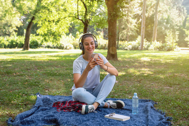 Joven chica hermosa en el parque con auriculares inalámbricos en la cabeza escuchando música y divirtiéndose con sonrisa en la cara - Foto, Imagen