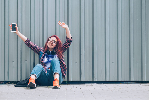 Young beautiful modern smiling female teenager in a checkered shirt and jeans with headphones and smartphone sitting near a wall on the street and Cheerful hands rising UP.Carefree student age concept - Φωτογραφία, εικόνα