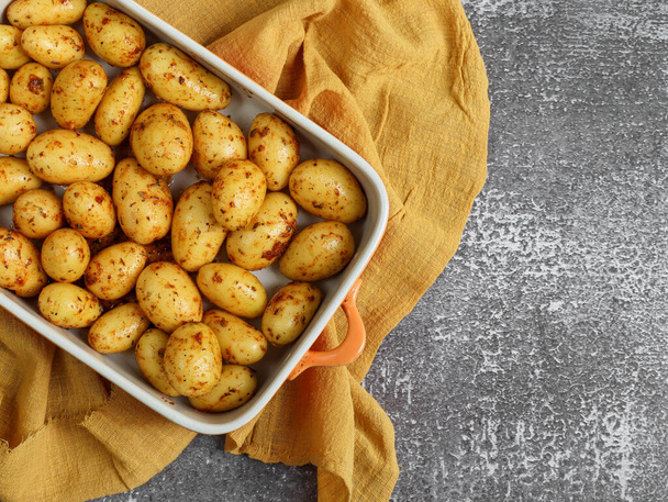 Potatoes with herbs and spicy paprika in an orange dish lie on a stone background with a yellow kitchen napkin on the left and a place for text on the right, top view. - Photo, Image