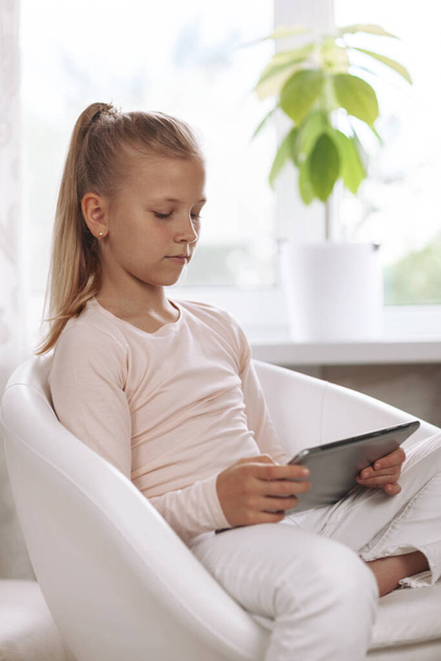Beyaz bir odada beyaz koltukta oturan güzel genç kız ödevlerini tablet, online eğitim ve uzaktan öğrenme kullanarak yapıyor., - Fotoğraf, Görsel