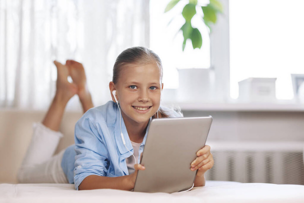 Adolescente blonde allongée sur le canapé joue à un jeu sur la tablette et communique par lien vidéo avec des amis. Technologies numériques, éducation en ligne - Photo, image