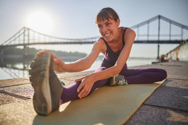 Een vooraanzicht van glimlachende jonge vrouw die doet Head-Knee Pose out yoga en is lachend outdoor - Foto, afbeelding