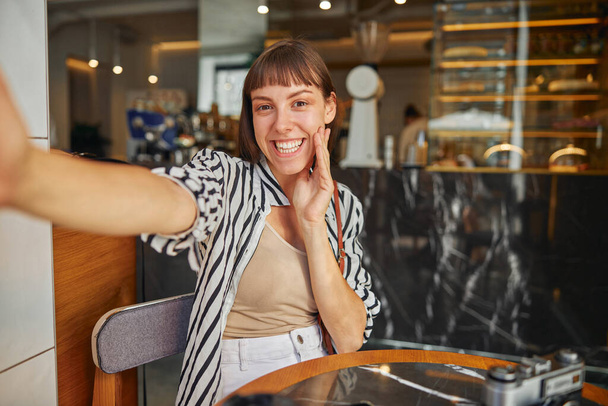 Cheerful young woman photographer  with outstretched hand in cafe close up - 写真・画像