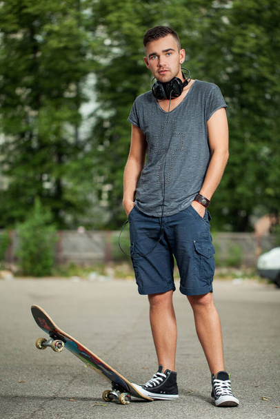Retrato de un chico guapo que está usando auriculares y de pie en su monopatín en un parque
 - Foto, Imagen