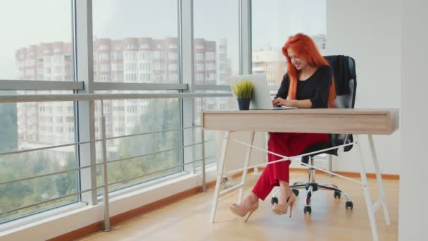 Fiatal vonzó nő vörös hajjal dolgozik egy fényes irodában, panorámás ablakokkal dolgozik egy laptop. - Felvétel, videó