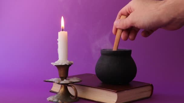 Czarna dzbanek czarownica jest palenie na starej książce i ręcznie mieszając z drewnianą łyżką, i świeca w świecznik spalania  - Materiał filmowy, wideo