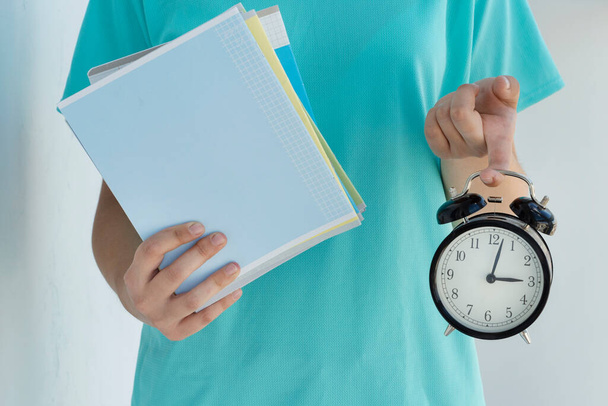 close up image of young female holds black alarm clock and stack of notebooks in her hands on light background. Copy space for text. Back to school concept, beginning of school year. Managing activities on time. - Photo, Image