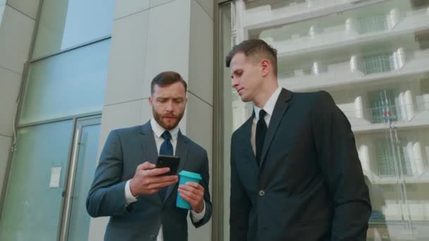 Dos empleados masculinos interactuando entre sí en el distrito de negocios del centro. Empresarios confiados que usan teléfonos inteligentes discutiendo la planificación de proyectos de negocios de computadoras en equipo corporativo - Imágenes, Vídeo