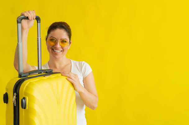 Smiling woman in sunglasses holding suitcase with retractable handle on yellow background - Foto, Bild