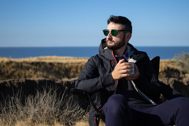 サングラスの若いブルネットの男とコーヒーの鉄マグカップカップを保持防水ジャケットは、海で崖の上のキャンプチェアに一人で座って、自然の風景の中で休暇を楽しんでいます。 - 写真・画像