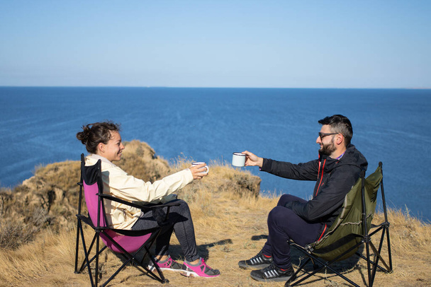 Reizen paar, jonge vrouw en man met kopjes koffie, zitten in stoelen ontspannen, doorbrengen herfstvakantie in het kamp in de buurt van de blauwe zee, genieten van een vakantie samen in de natuur landschap. - Foto, afbeelding