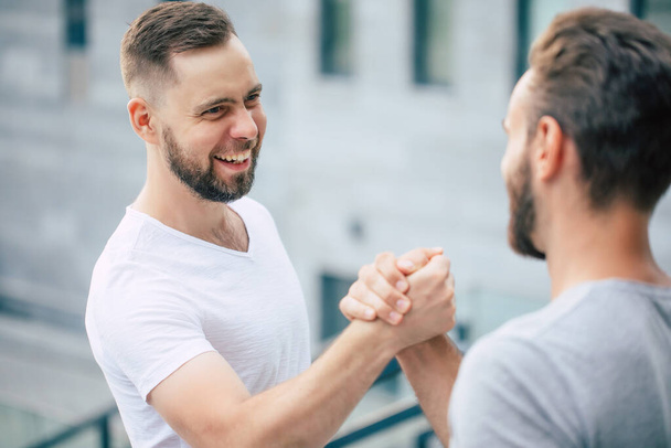 Men's friendly handshake. Two young bearded handsome friends in casual t-shirts are posing together on the city background.  - Photo, image