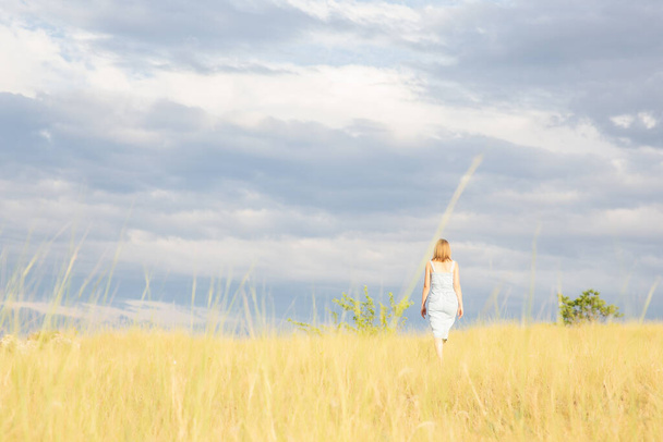 Uma menina com longos cabelos loiros caminha através de um campo de grama amarela seca contra um belo céu - Foto, Imagem