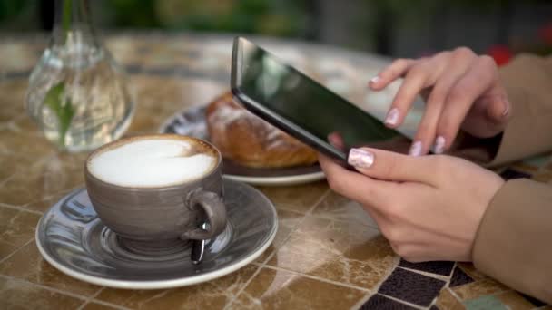Jovem mulher de negócios senta-se em um café e escreve mensagens de texto em um tablet close-up. Menina com café e um pão na varanda de verão. - Filmagem, Vídeo