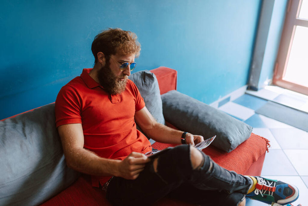 Un giovane caucasico con una maglietta rossa è seduto in sala d'attesa dal parrucchiere e sta leggendo una rivista - Foto, immagini