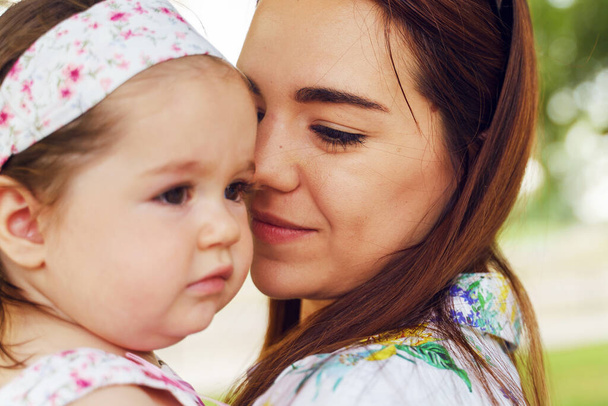 Close up of young caucasian woman holding her one year old child in park - Mother and little daughter standing outdoor in the city in summer day bonding - motherhood and growing up concept - Photo, Image