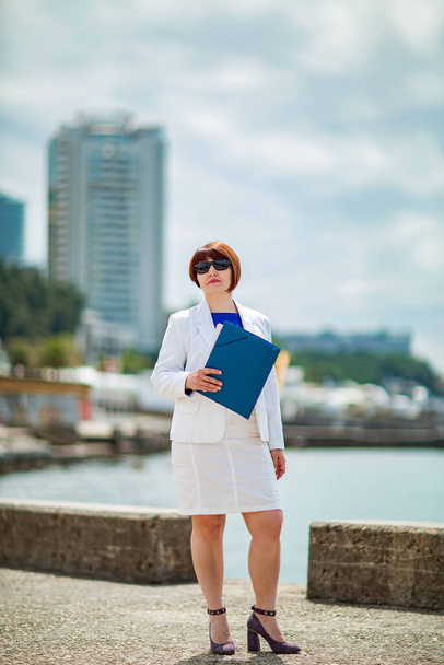 A beautiful woman, brown-haired, in a white business suit and blue blouse,with documents in her hands, stands against the background of the city near the sea, a Business portrait . - Fotoğraf, Görsel