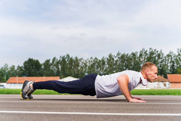 Low angle side view on adult caucasian man doing push-ups on the running track - Blonde male athlete in stadium training in summer day - real people healthy lifestyle concept full length - Photo, Image