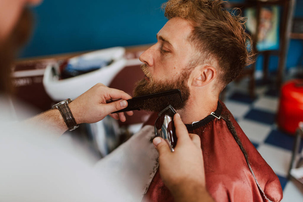 Gros plan des mains d'un coiffeur taillant la barbe d'un jeune homme caucasien. Nouvelle barbe et coiffure - Photo, image