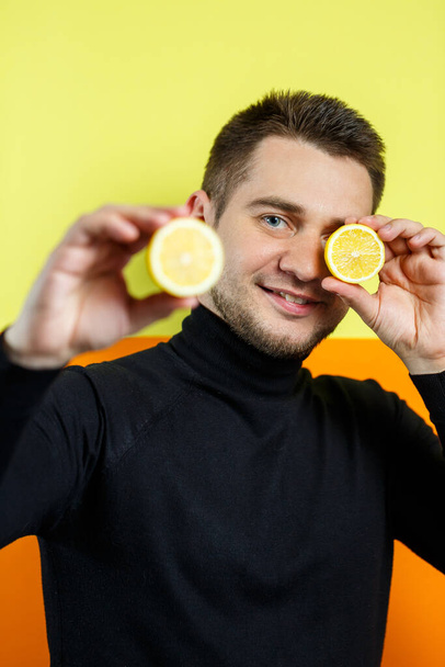 Portrait of an elegant man on a yellow background in black raglan with a sliced lemon in his hands. Stylish guy. A smile on his face. An image for a male photo. Photo session concept - Foto, Imagen