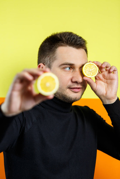 Porträt eines eleganten Mannes auf gelbem Hintergrund in schwarzem Raglan mit einer Zitronenscheibe in den Händen. Stylischer Typ. Ein Lächeln auf seinem Gesicht. Ein Bild für ein Männerfoto. Konzept für Fototermine - Foto, Bild