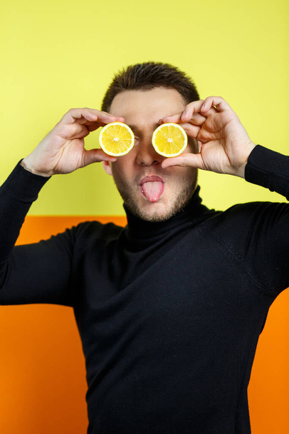Портрет человека на жёлтом фоне в чёрном раглане с вырезанным лимоном вместо глаз. Парень с лимонами у лица. Улыбка на его лице. Изображение для мужского фото. Концепция фотосессии - Фото, изображение